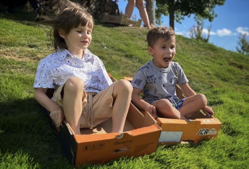 Viagem em Família: duas crianças escorregando na grama dentro de caixas de papelão